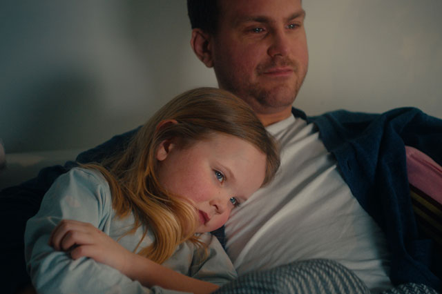 Pappa och dotter sitter tätt intill varandra i en soffa och tittar på något tillsammans.